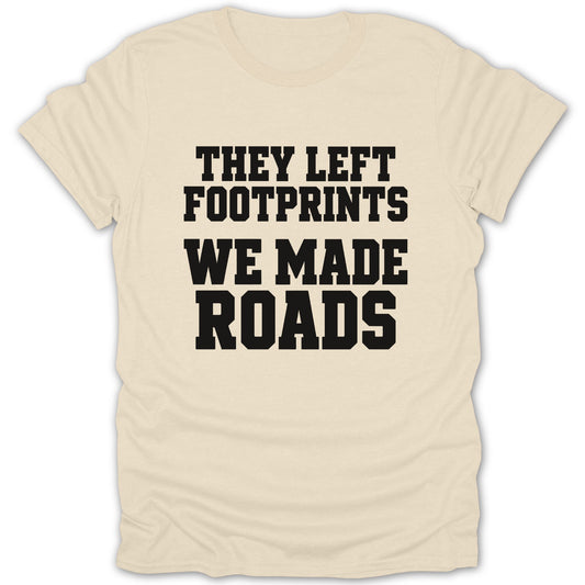 A cream-colored short-sleeved t-shirt featuring black block lettering that reads THEY LEFT FOOTPRINTS WE MADE ROADS.