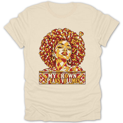 A cream-colored t-shirt featuring a stained glass style graphic of a Black woman with an afro and the text "MY CROWN SPEAKS VOLUMES".
