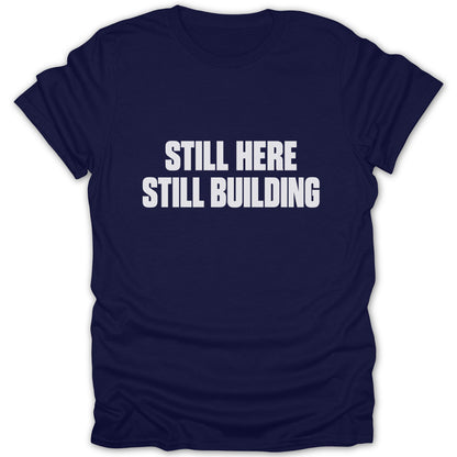 Navy blue short-sleeved t-shirt with white block text reading STILL HERE STILL BUILDING on the chest.