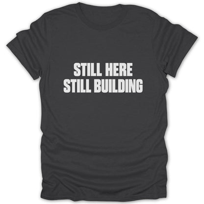 A dark gray t-shirt with white text reading "STILL HERE STILL BUILDING" centered on the chest.