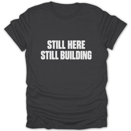 A dark gray t-shirt with white text reading "STILL HERE STILL BUILDING" centered on the chest.