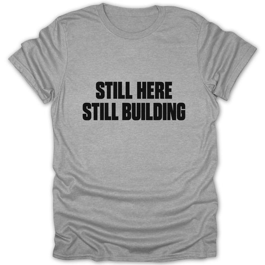 A grey short-sleeved t-shirt with the black text STILL HERE STILL BUILDING centered on the chest.