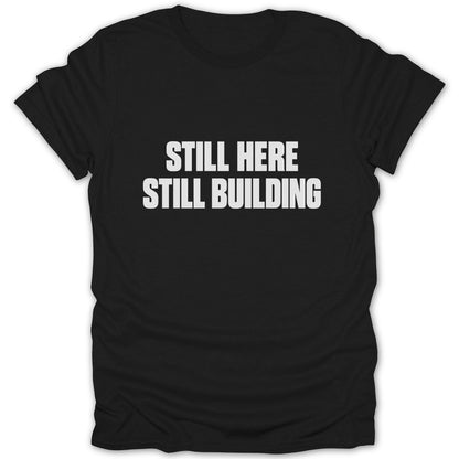 A black short-sleeved t-shirt with white block text on the chest that reads STILL HERE STILL BUILDING.