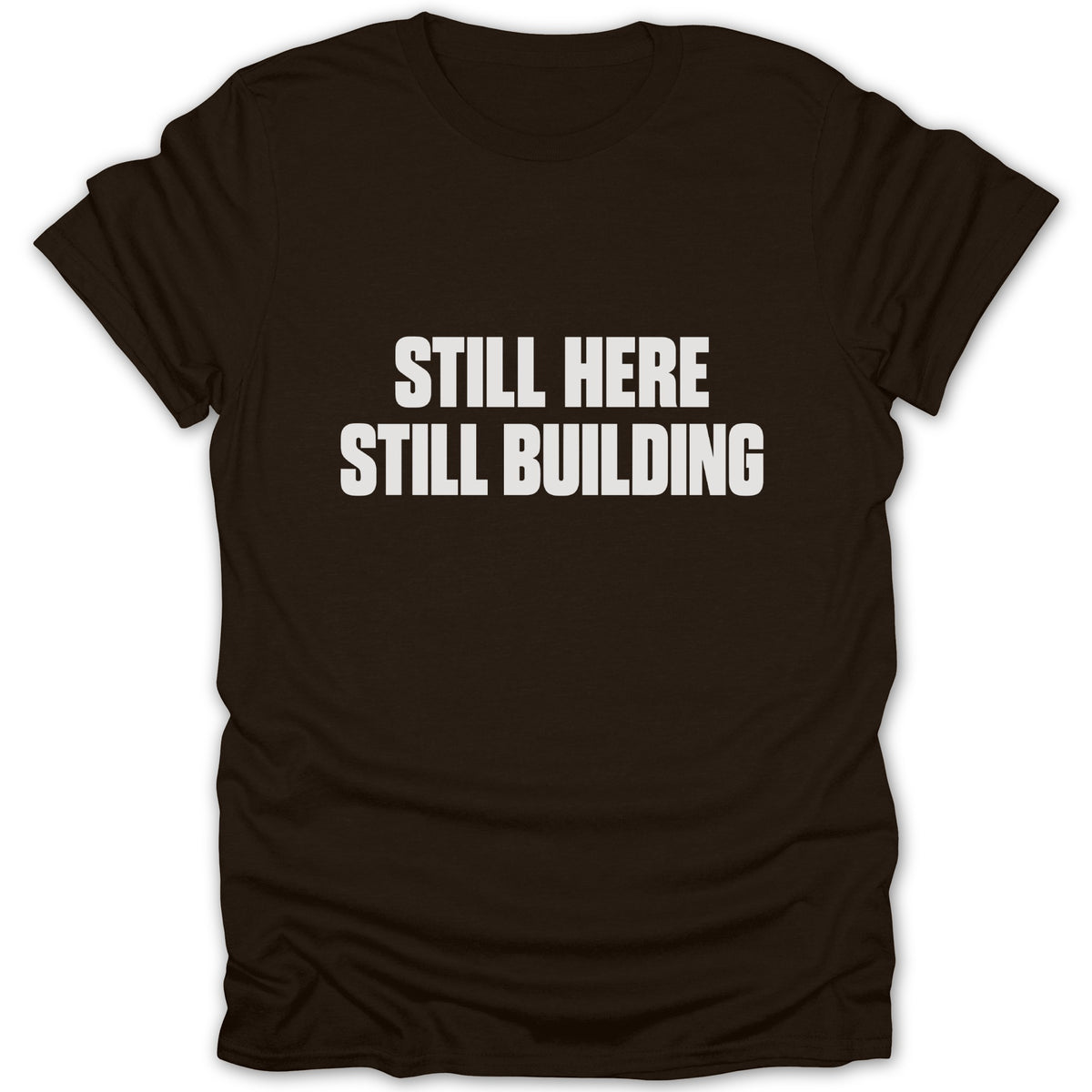 A dark brown short-sleeved t-shirt featuring the white text STILL HERE STILL BUILDING centered on the chest.