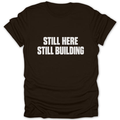 A dark brown short-sleeved t-shirt featuring the white text STILL HERE STILL BUILDING centered on the chest.