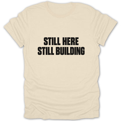 Cream crewneck t-shirt with black text on the chest that reads "STILL HERE STILL BUILDING."