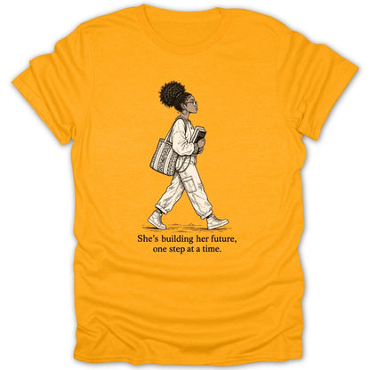 Yellow t-shirt with graphic of woman walking, holding books, text: "She's building her future, one step at a time."
