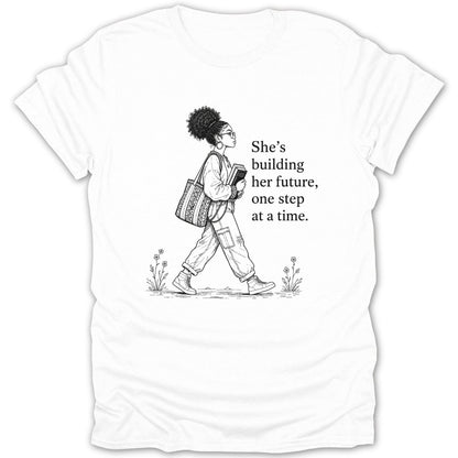 Black and white graphic on white t-shirt of a woman carrying books, with text "She's building her future, one step at a time."