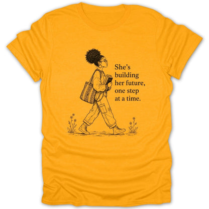Yellow t-shirt with drawing of woman walking, holding books, and text "She's building her future, one step at a time."