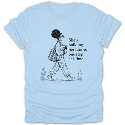 Light blue t-shirt with a sketch of a woman walking and text "She's building her future, one step at a time."